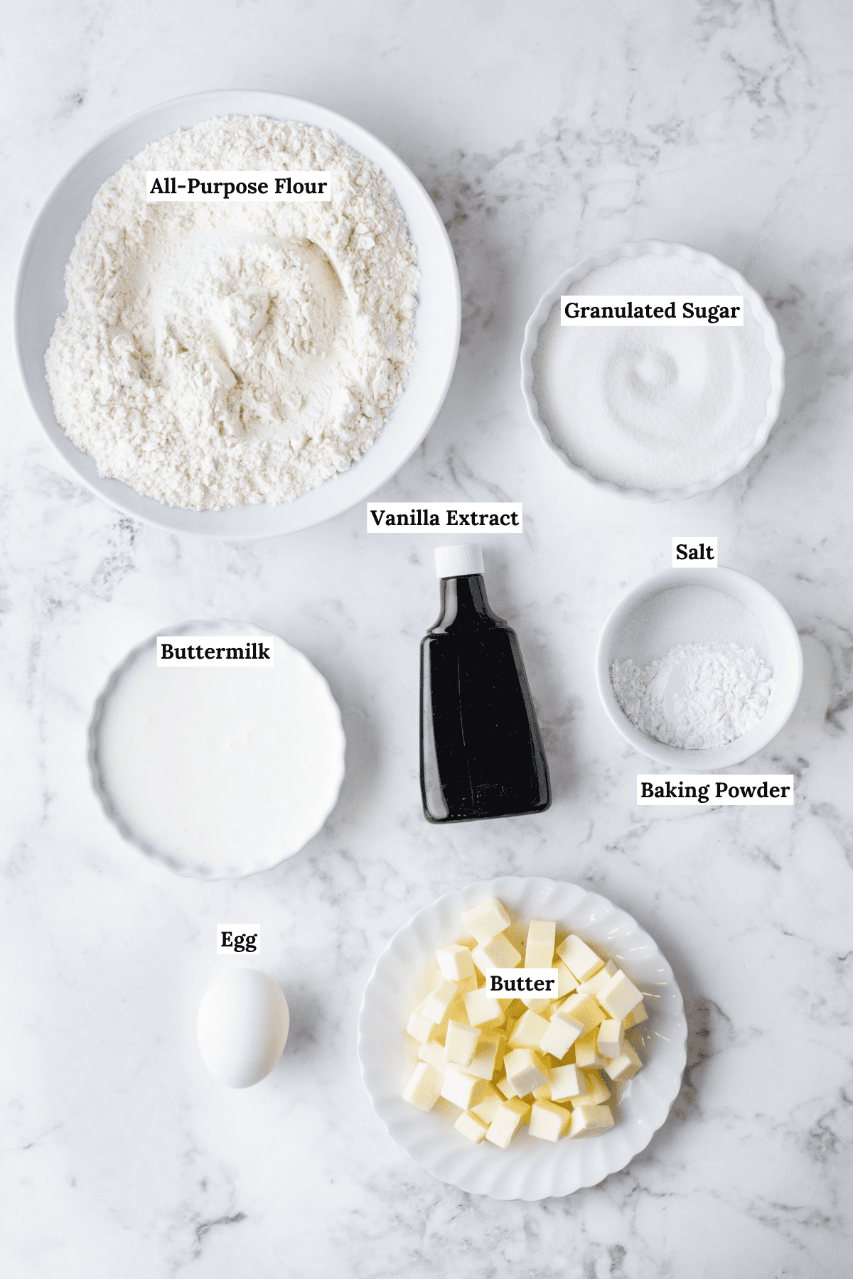 over head view of the ingredients for scones including a bowl of all-purpose flour, granulated sugar, buttermilk, salt, vanilla extract, baking powder, an egg and a plate of cubed butter
