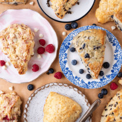 Overhead view of assorted scones on decorative plates, surrounded by fresh berries and white chocolate chips. A knife rests on one plate. Text at the top reads Master Scone Recipe, highlighting the perfect way to enjoy homemade scones.