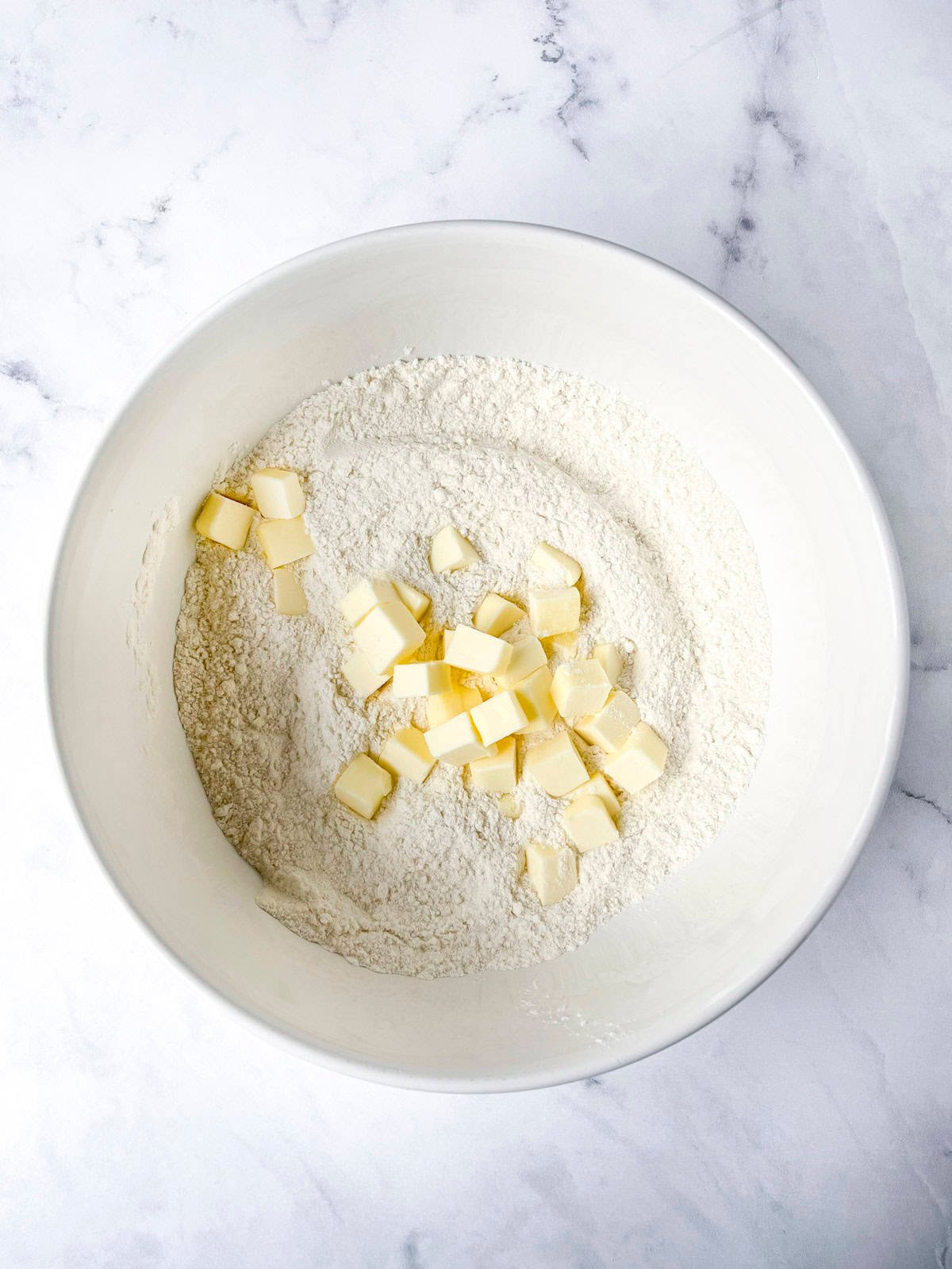 A white bowl containing flour and small cubes of butter on a marble surface, ready for mixing.